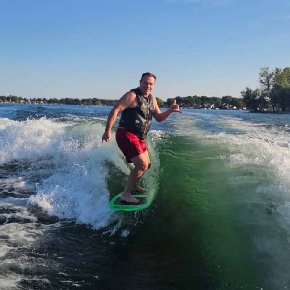 John Tremaine wakesurfing on Lake Fenton on a sunny day, giving a shaka sign